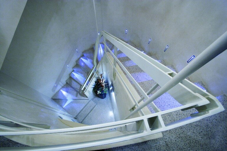 Scala elicoidale in pietra chiara fotografata dall'alto, con illuminazione incassata nei gradini